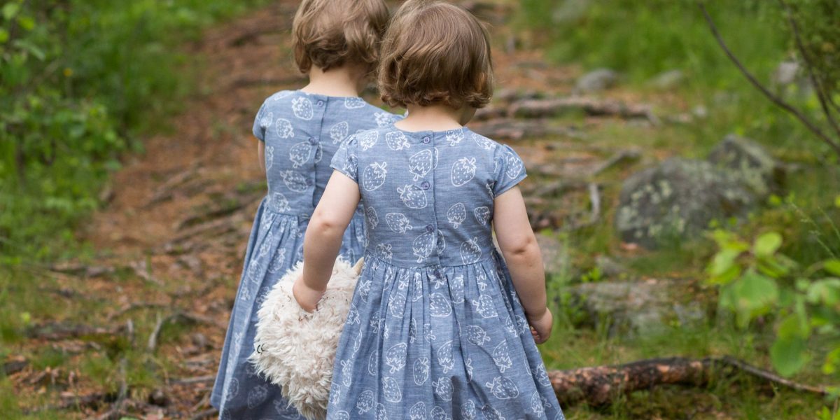 pienet kaksostytöt, selin, kävelevät samanlaiset siniharmaat mekot päällä pitkin kesäistä metsäpolkua.
