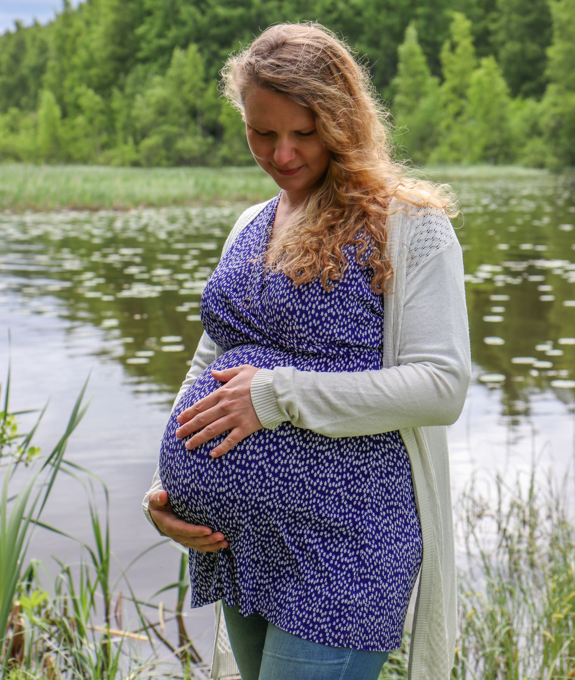 kaksosia odottava äiti kesäisessä järvimaisemassa kädet vatsan ympärillä.