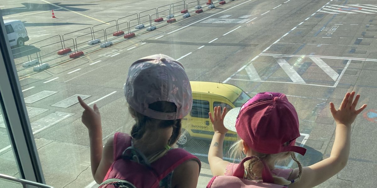 kaksostytöt katselevat lentokentällä ikkunasta ulos kiitoradalle.