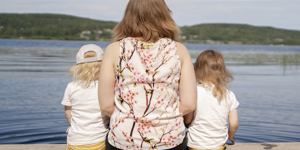 Äiti ja kaksi lasta istuvat laiturilla kasvot järvelle päin.