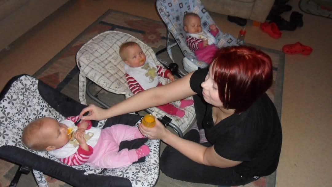 Äiti istuu lattialla syöttämässä sosetta kolmessa sitterissä istuville kolmosvauvoilleen.