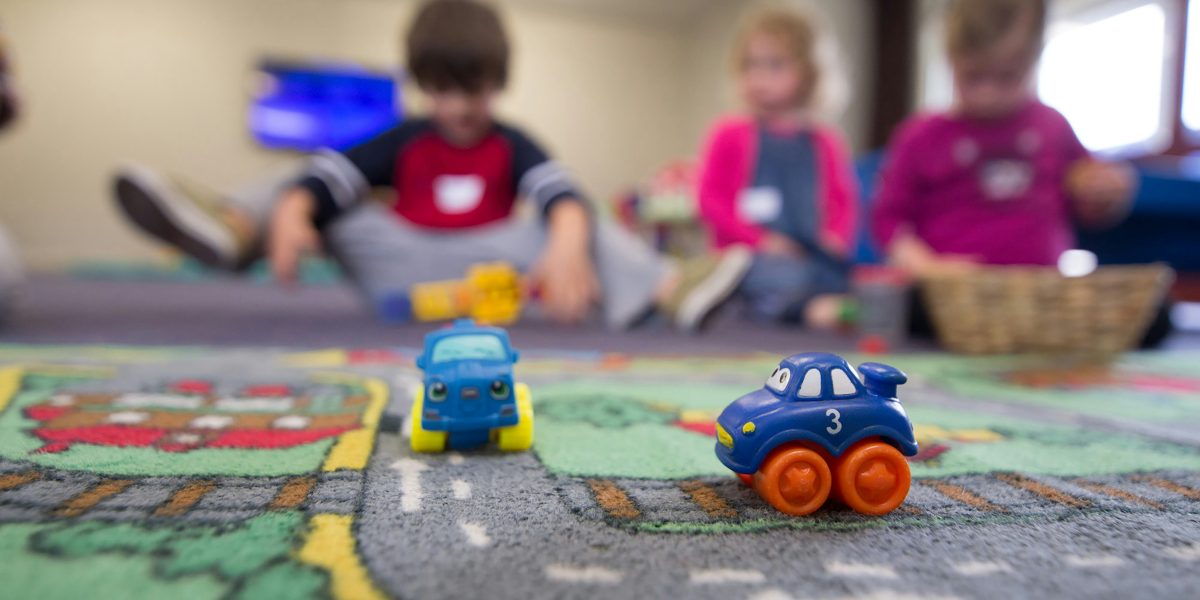 Etualalla lasten liikennematto, jonka päällä on kaksi sinistä leluautoa. Taustalla on kolme lattialla leikkivää lasta.