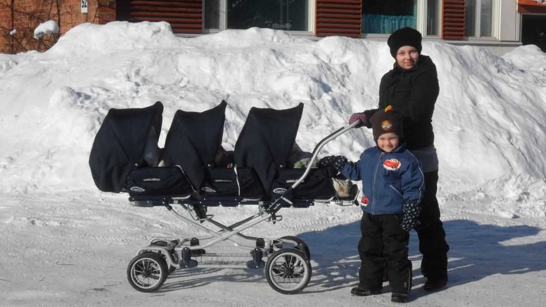 Äiti ja isosisarus ulkoiluttamassa kolmosvauvoja kolmosten rattaissa aurinkoisessa talvisäässä.