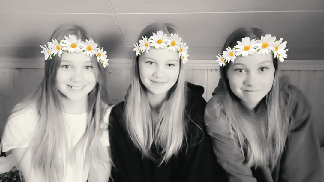 Teini-ikäiset kolmossisarukset istuvat rivissä mustavalkokuvassa valokuvafiltterillä lisätyt kukkaseppeleet päässään.
