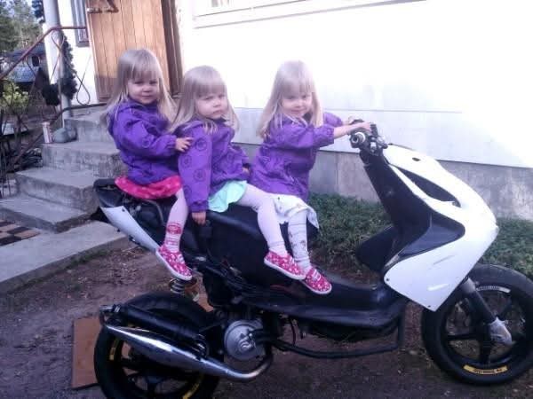 Kolmossisarukset istuvat kaikki yhden skootterin päällä. Lapsilla on yllään violetit tuulitakit, eriväriset hameet sekä pinkit sukkahousut ja tennarit.