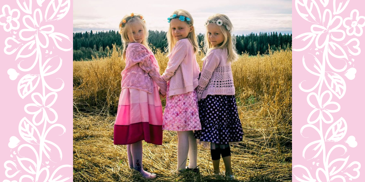 Kolmossisarukset kukkaseppeleet päässä viljapellolla. Lapset seisovat rivissä ja pitävät toisiaan kädestä olkansa yli kohti kameraa kurkistaen vaaleanpunaiset neuletakit ja mekot yllään.