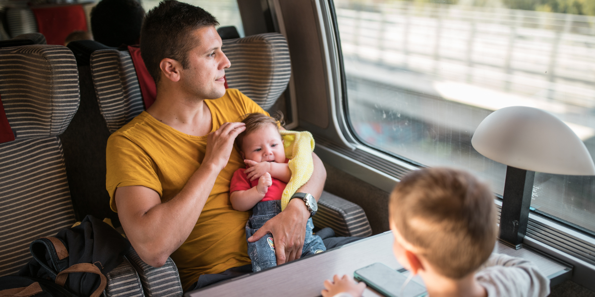 Valkoinen isä lapsineen junassa. Isä istuu ikkunapaikalla vauva sylissään ja katselee ulos ikkunasta. Etualalla selin kameraan on lapsi, joka nojaa käsillään hänen ja muun perheen välissä olevaan pöytään.