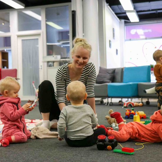 Äidit ja lapset leikkivät ja seurustelevat monikkokahvilatapahtumassa, toiset sohvilla istuen ja toiset lattialla.