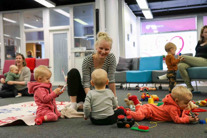 Äidit ja lapset leikkivät ja seurustelevat monikkokahvilatapahtumassa, toiset sohvilla istuen ja toiset lattialla.