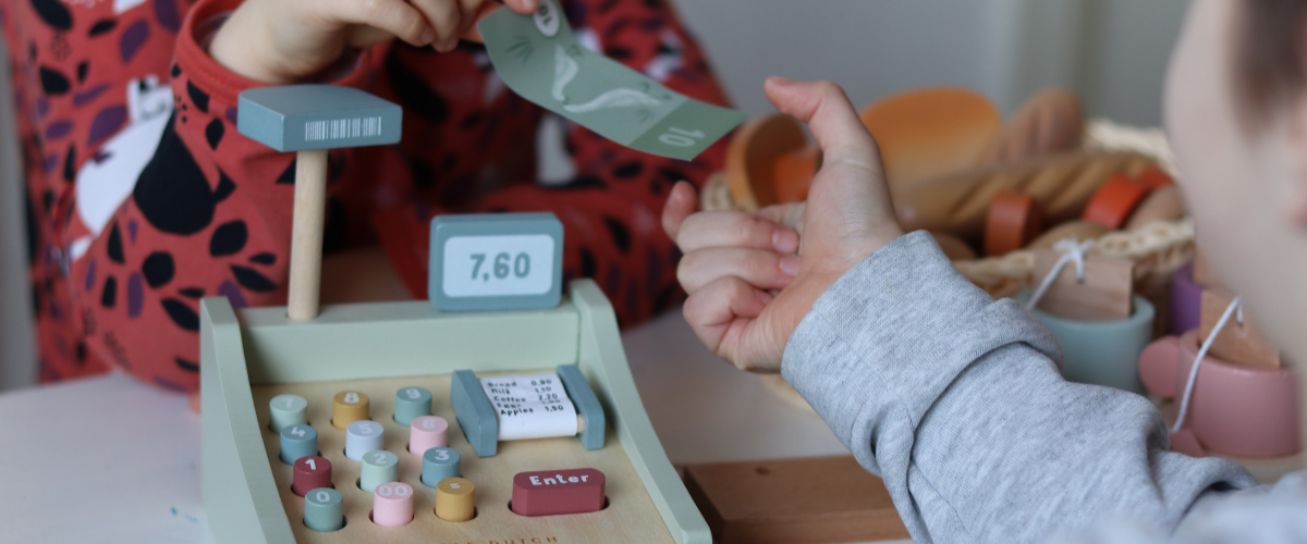 Lapsi luovuttaa leikkirahaa toiselle lapselle leikkikassakoneen ylitse.