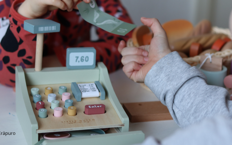 Lapsi luovuttaa leikkirahaa toiselle lapselle leikkikassakoneen ylitse.