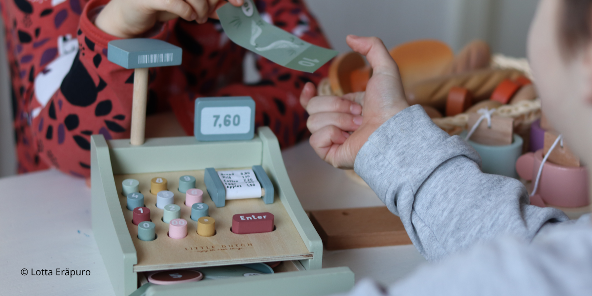 Lapsi luovuttaa leikkirahaa toiselle lapselle leikkikassakoneen ylitse.