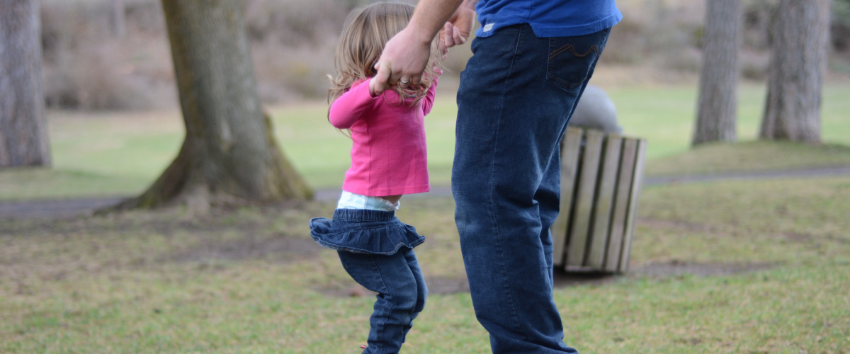 Kuvassa näkyy aikuisen jalat ja kädet, jotka pitävät kiinni pientä tyttöä hänen käsistään, kun hän hyppii trampoliinilla.