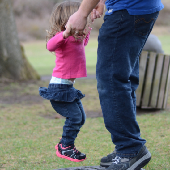 Kuvassa näkyy aikuisen jalat ja kädet, jotka pitävät kiinni pientä tyttöä hänen käsistään, kun hän hyppii trampoliinilla.
