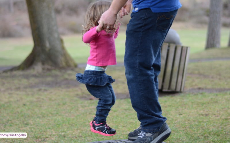 Kuvassa näkyy aikuisen jalat ja kädet, jotka pitävät kiinni pientä tyttöä hänen käsistään, kun hän hyppii trampoliinilla.