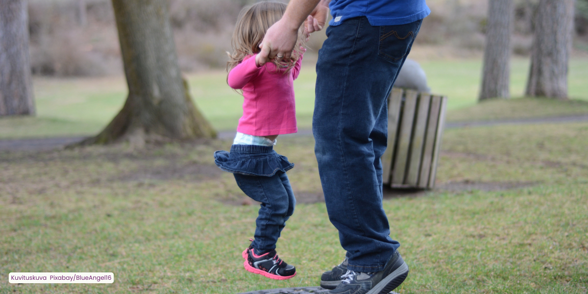 Kuvassa näkyy aikuisen jalat ja kädet, jotka pitävät kiinni pientä tyttöä hänen käsistään, kun hän hyppii trampoliinilla.