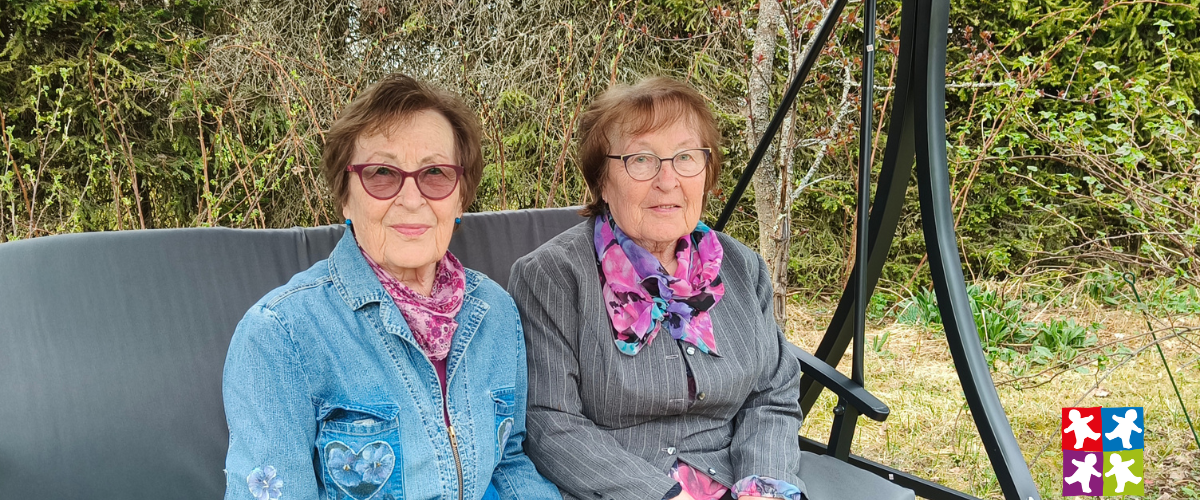 80-vuotiaat kaksossiskoset istuvat ulkona puutarhakeinussa vierekkäin.