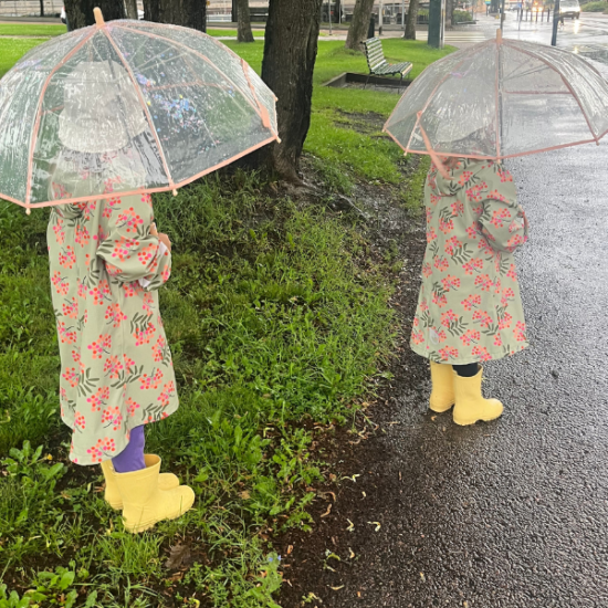 Kaksi tyttöä seisoo selin puiston laidalla sadetakki päällä ja kumisaappaat jalassa sateenvarjojaan pidellen.