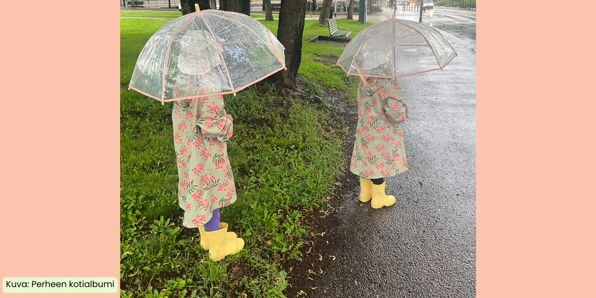 Kaksi tyttöä seisoo selin puiston laidalla sadetakki päällä ja kumisaappaat jalassa sateenvarjojaan pidellen.