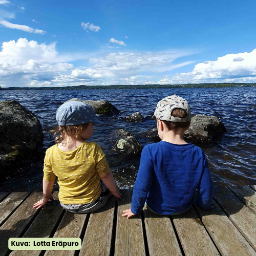 Kaksi pientä lasta istuu laiturilla, lippikset päässä, aurinkoisessa säässä meren rannalla ja taivas on sininen ja siellä on valkoisia pilviä.